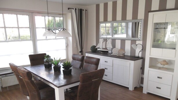 A dining area with a wooden table, six chairs, and potted plants. There is a white cabinet with drawers and a mirror above it. - Haferweg 2, Strandnelke EG - Haferweg 2, Strandnelke (Wyk auf Föhr)