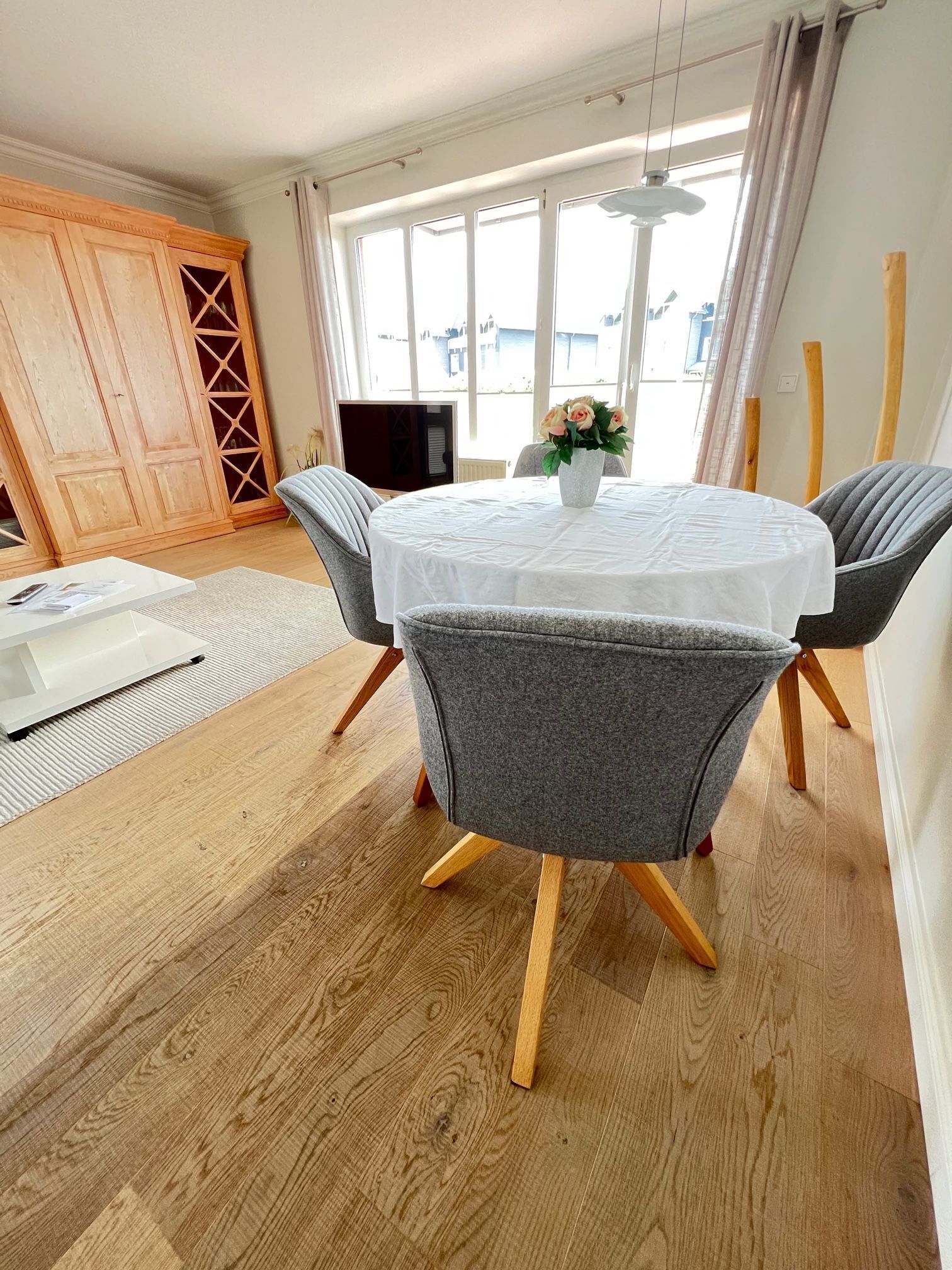 Un coin repas avec une table ronde, des chaises à revêtement gris, une nappe blanche et un vase rempli de fleurs.