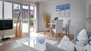 A modern living room with a sofa, coffee table, and a TV. There is a dining area with chairs and a table, and a balcony with a view of buildings. - Beach time, Germany (List)