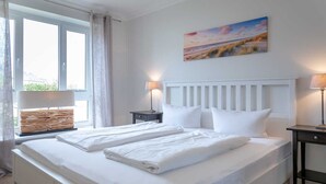 A neatly made double bed with white linens, a headboard, two bedside tables with lamps, and a window with curtains. - Beach time, Germany (List)