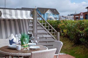 A wooden deck with a table set for dining, surrounded by wicker chairs. A striped awning provides shade. - Beach time, Germany (List)