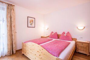 A bedroom with a wooden bed, checkered bedding, two bedside tables, a nightstand, a framed picture on the wall, and a window with curtains.