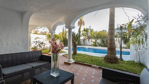 Uma área de estar coberta ao ar livre com vista para a piscina, um vaso com flores e um candeeiro junto à piscina.