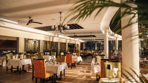 A spacious restaurant with multiple tables and chairs, ceiling fans, and a large window providing natural light. - Chobe Safari Lodge (Kasane)