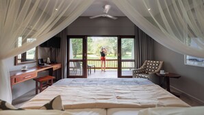 A bedroom with a large bed, a desk with a computer, a chair, and a view of a balcony with a person standing. - Chobe Safari Lodge (Kasane)