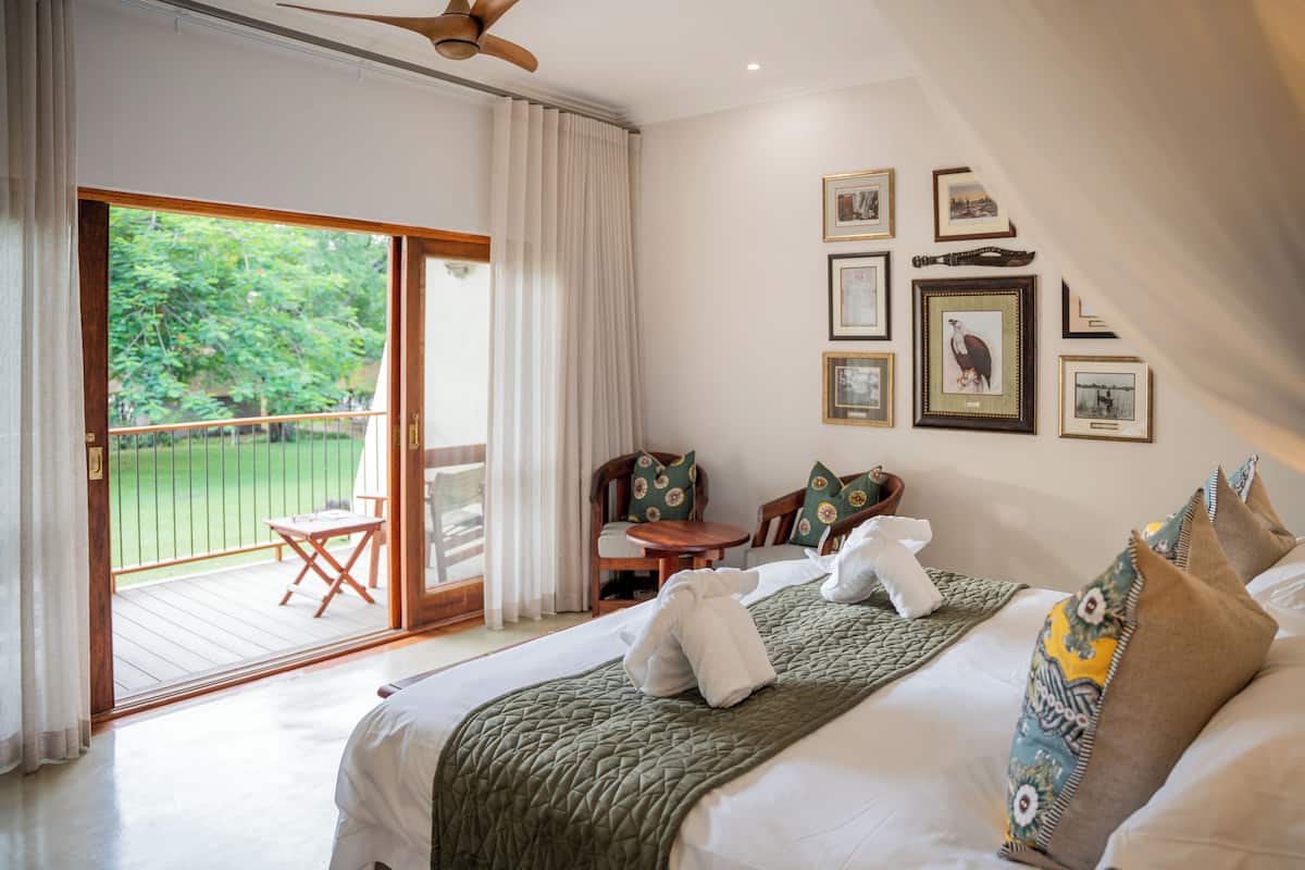 A hotel room with a large bed, a balcony with a view of the greenery, and a wall decorated with framed pictures.