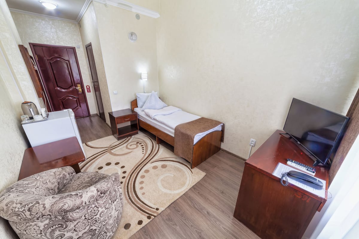 A compact hotel room with a bed, a desk with a TV, a small table with a lamp, and a patterned sofa.