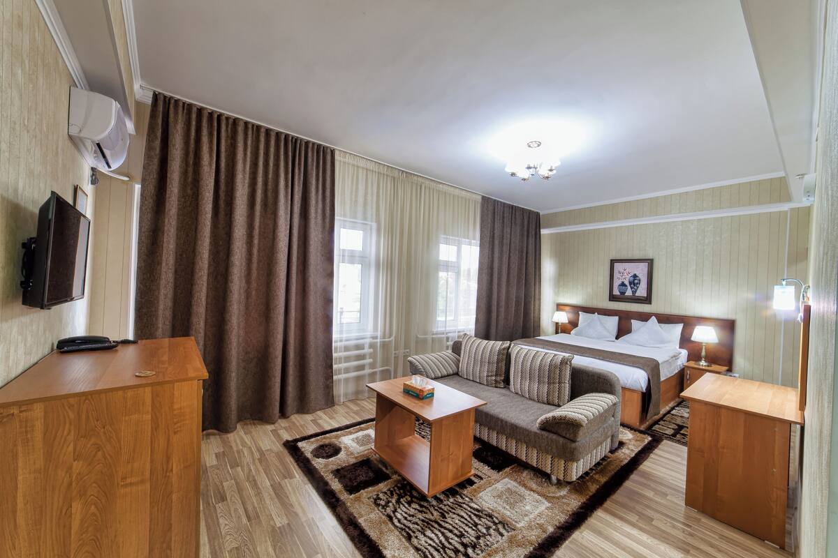 A hotel room with a bed, a sofa, a wooden side table, a television, and a window with curtains.