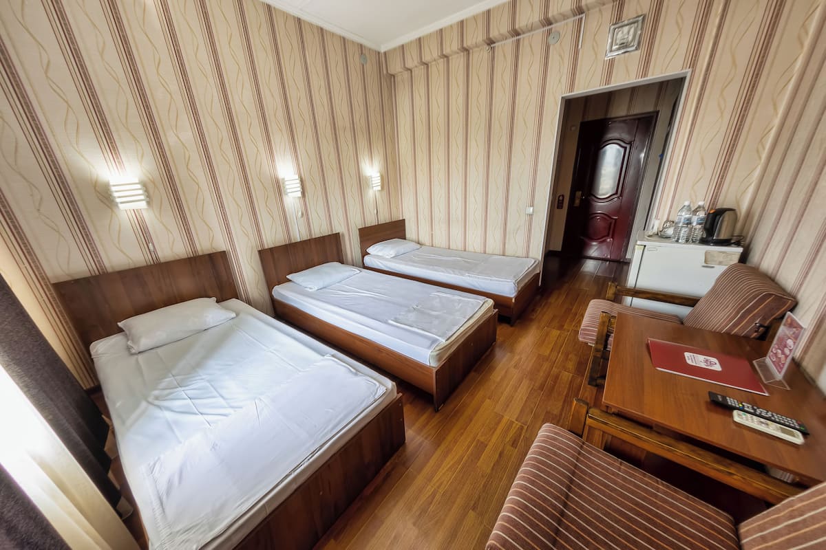A hotel room with three single beds, a wooden table, and a small desk with a red folder and remote controls.
