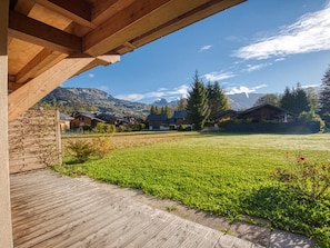 Une terrasse en bois qui mène à une zone herbeuse, avec des maisons en arrière-plan et des montagnes à l’horizon.
