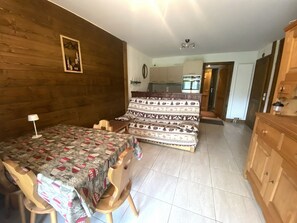 A cozy room with a bed, a wooden headboard, a small table with a lamp, and a view into the kitchen.