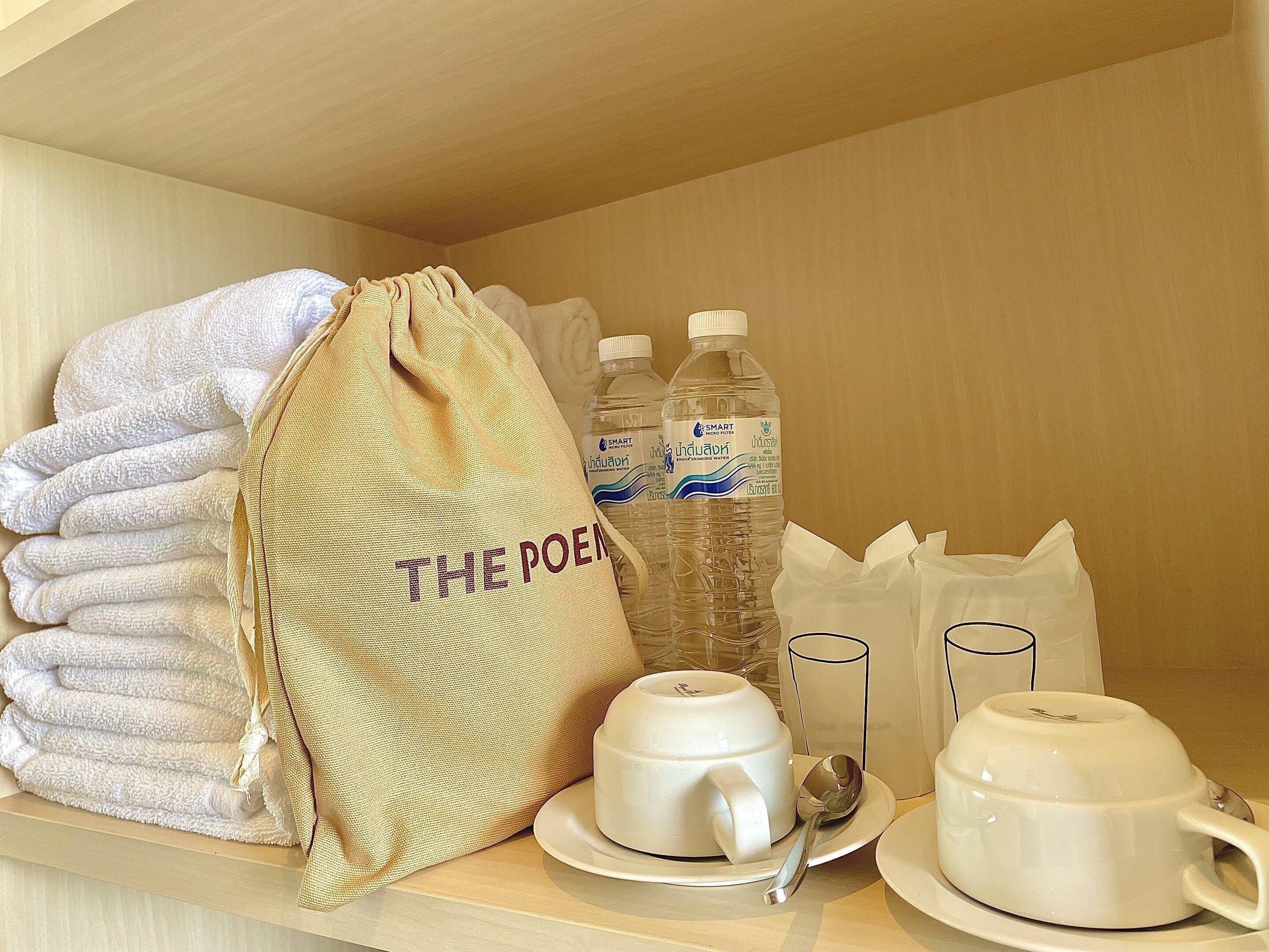 A neatly arranged hotel room with a stack of white towels, a beige bag labeled "THE POEM", two bottles of water, and a set of white cups and saucers.