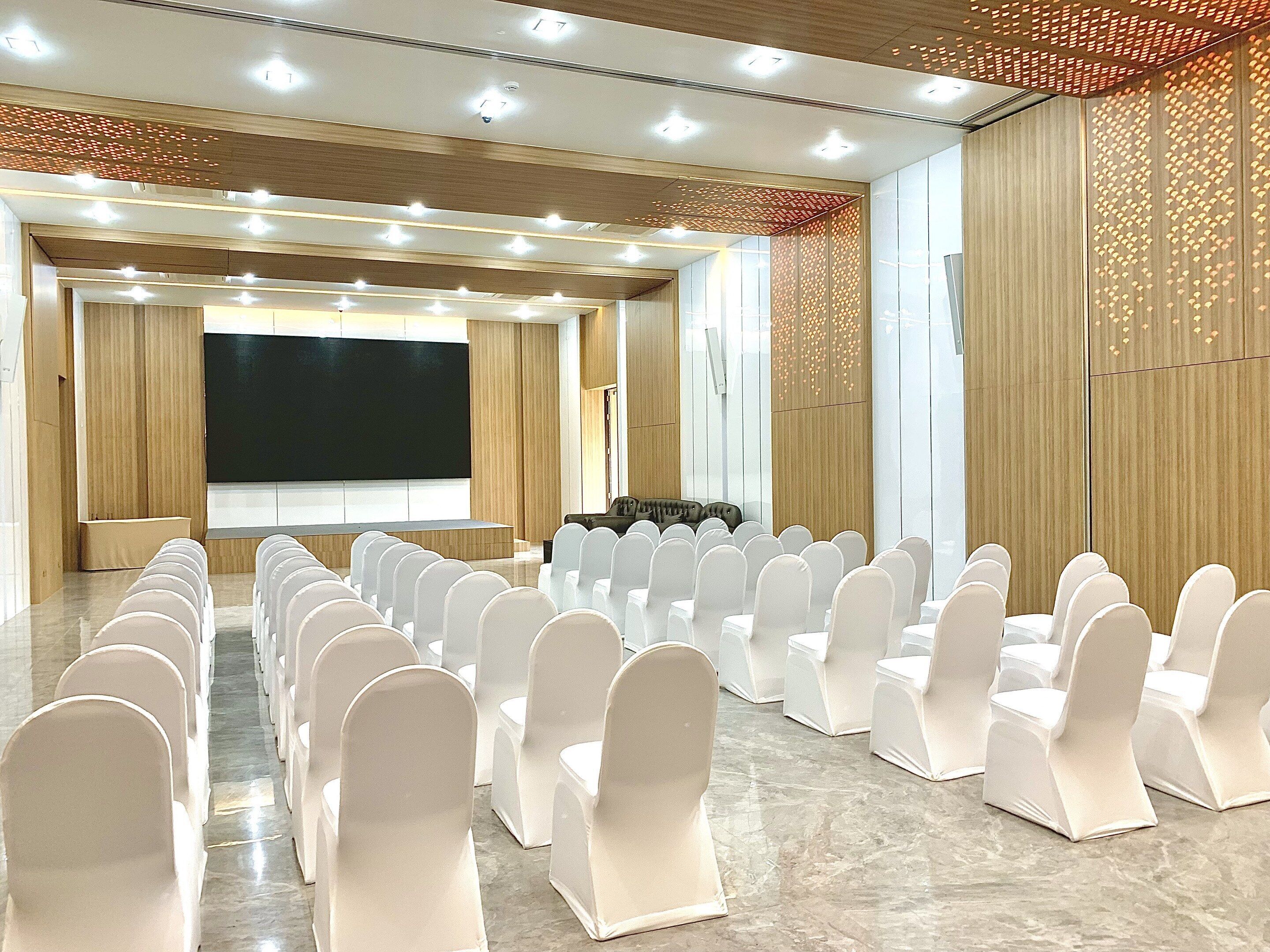 Un salón de conferencias con filas de sillas blancas, una pantalla grande y una zona para ponentes.