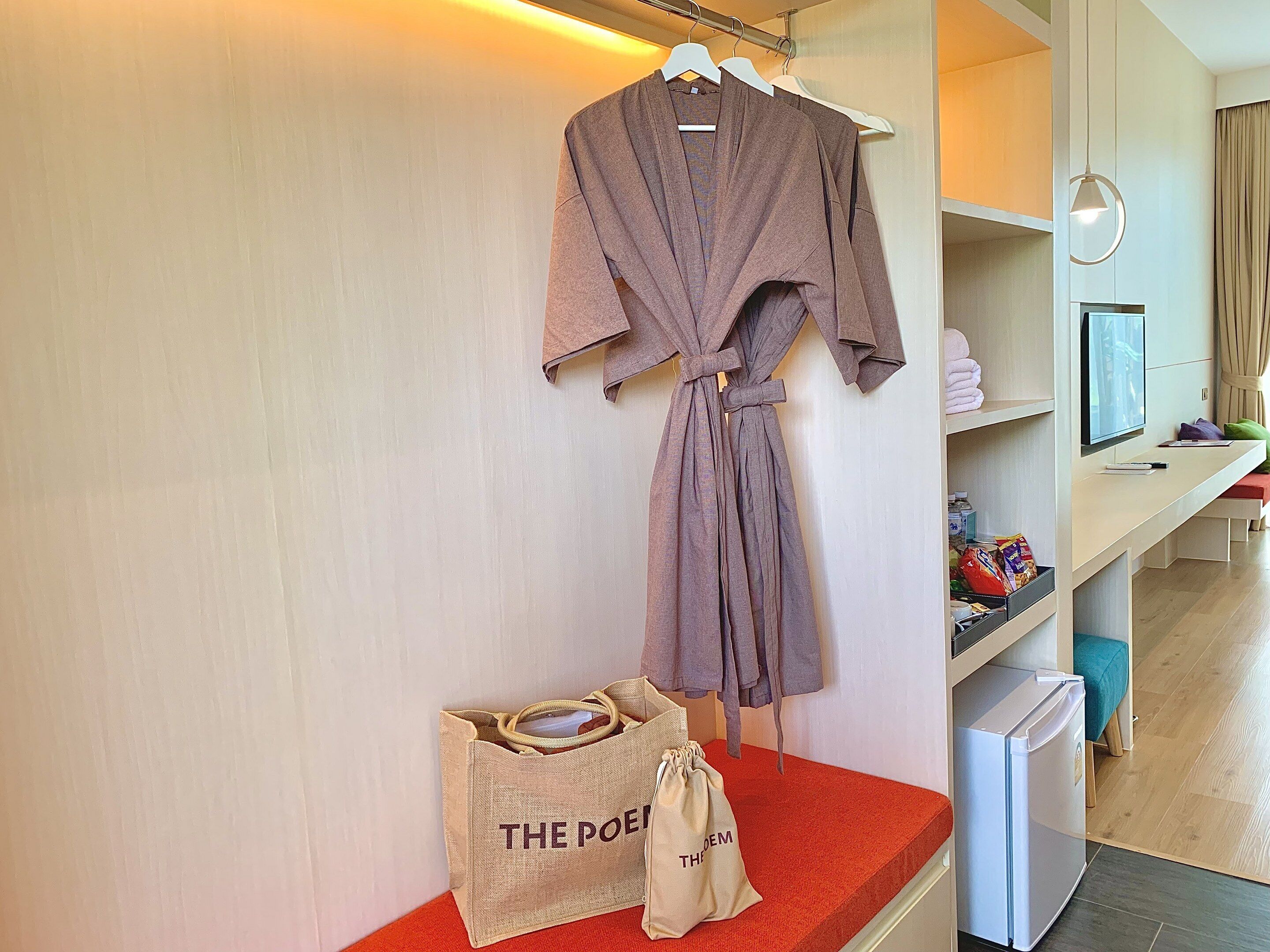 A robe hanging on a rack, a small bag labeled "THE POEM", and a mini fridge on a red platform in a hotel room.