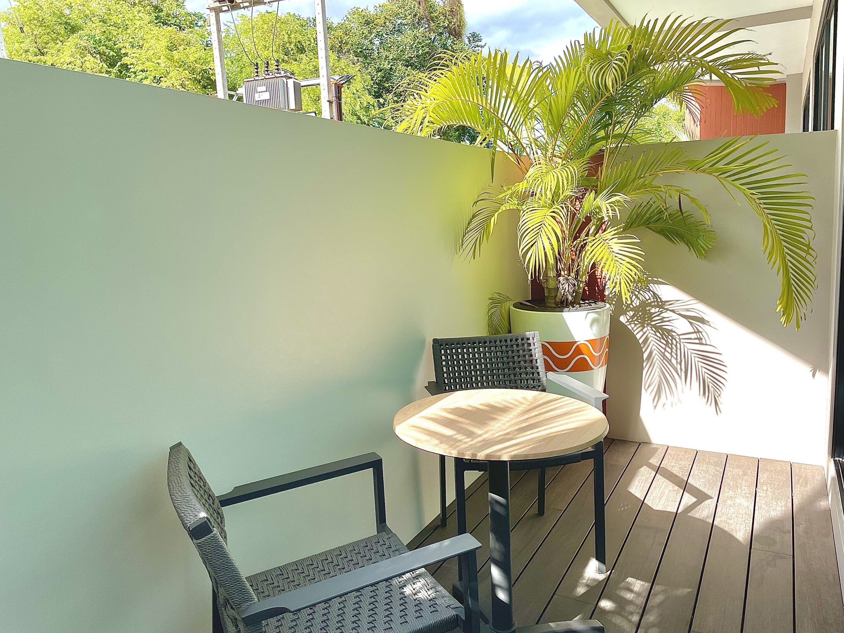 A balcony with a wooden table and two chairs, a potted plant, and a view of trees and a building in the background.