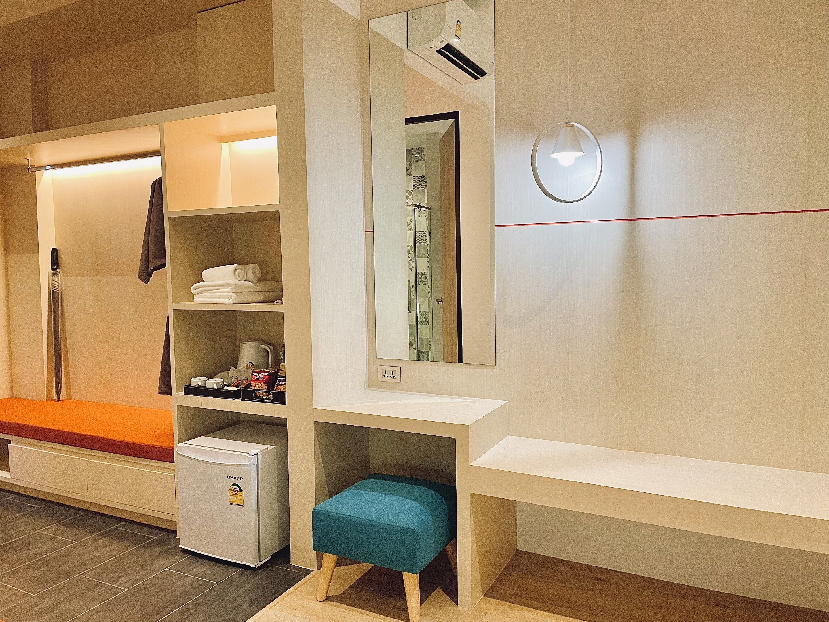 Habitación de hotel con zona de ducha, un pequeño banco, refrigerador compacto y espejo con marco decorativo.