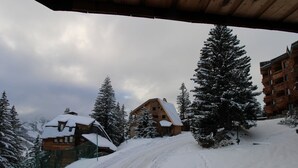 Une route de montagne enneigée menant à un chalet en bois au toit pentu, entouré d’arbres chargés de neige et d’un bâtiment à la façade rouge.