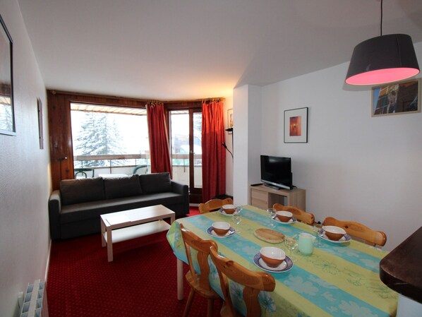 A cozy living room with a sofa, a dining table set for four, a television, and a balcony with red curtains.