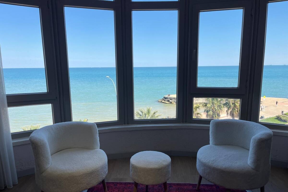 Une pièce avec vue sur la baie, deux chaises blanches, un petit pouf et un tapis rouge à motifs.