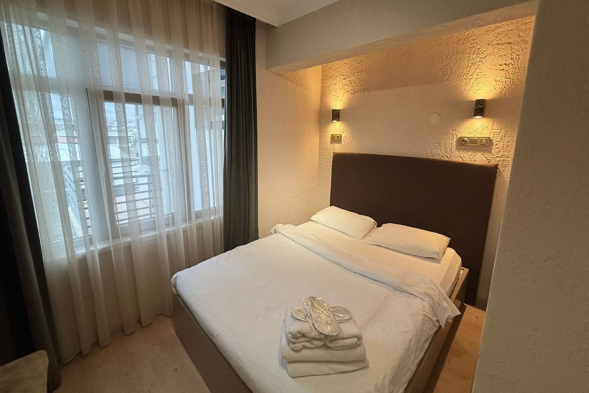 Une chambre d’hôtel avec un lit, une fenêtre avec des rideaux légers et deux lampes fixées au mur.