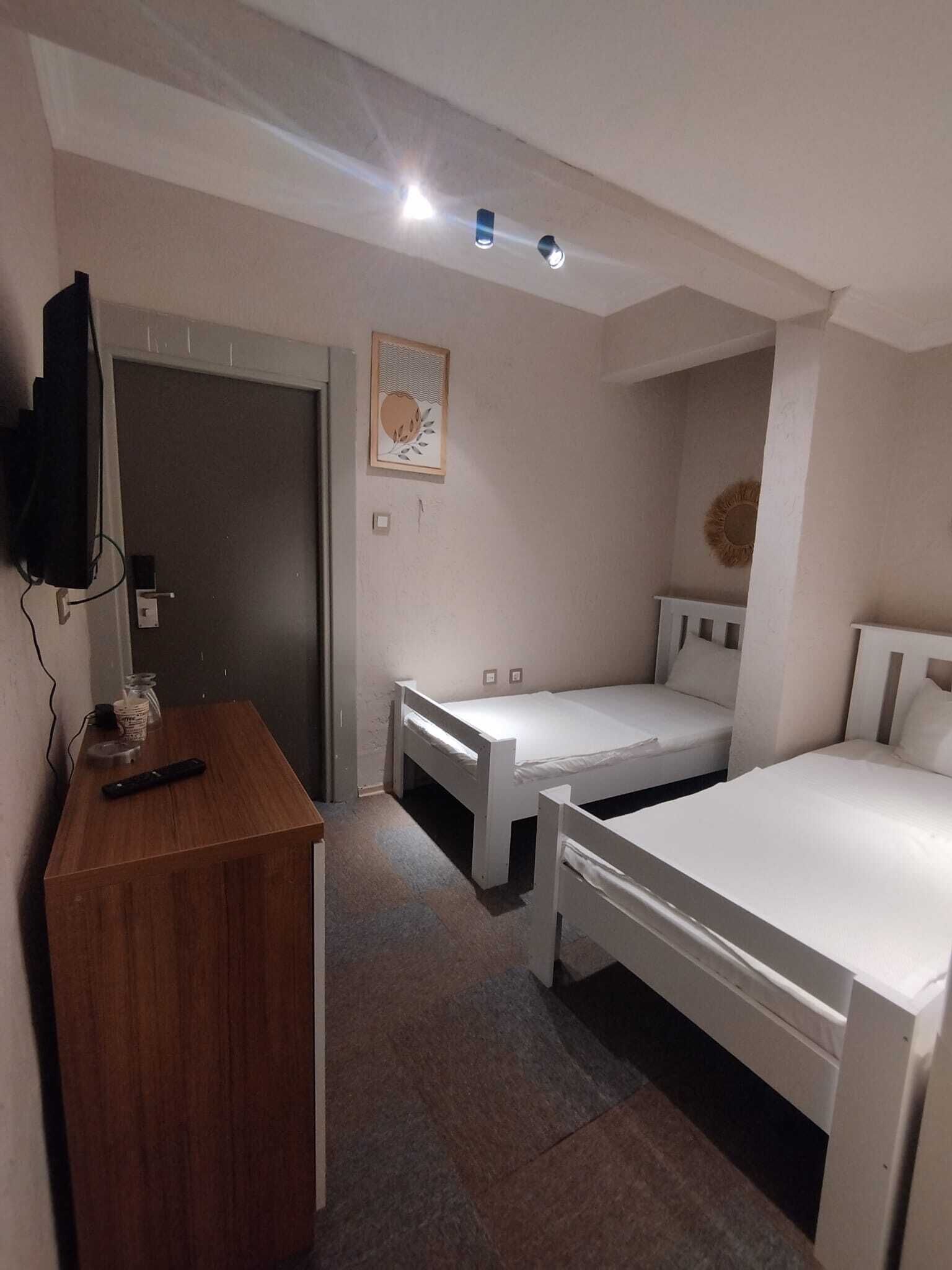 Une petite pièce, de taille modeste, avec un bureau en bois, deux lits simples, un téléviseur fixé au mur et un luminaire au plafond.