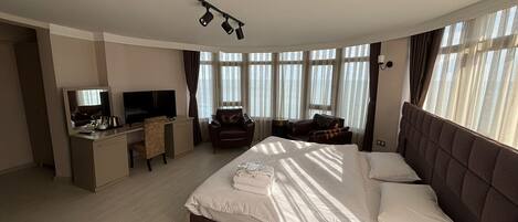 A modern hotel room with a large bed, a desk with a TV, and a view of the sea through large windows.