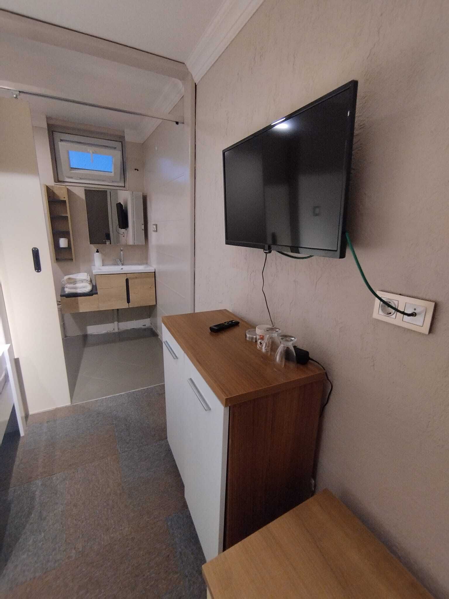 A compact hotel room with a kitchenette, a flat-screen TV mounted on the wall, and a small wooden table.