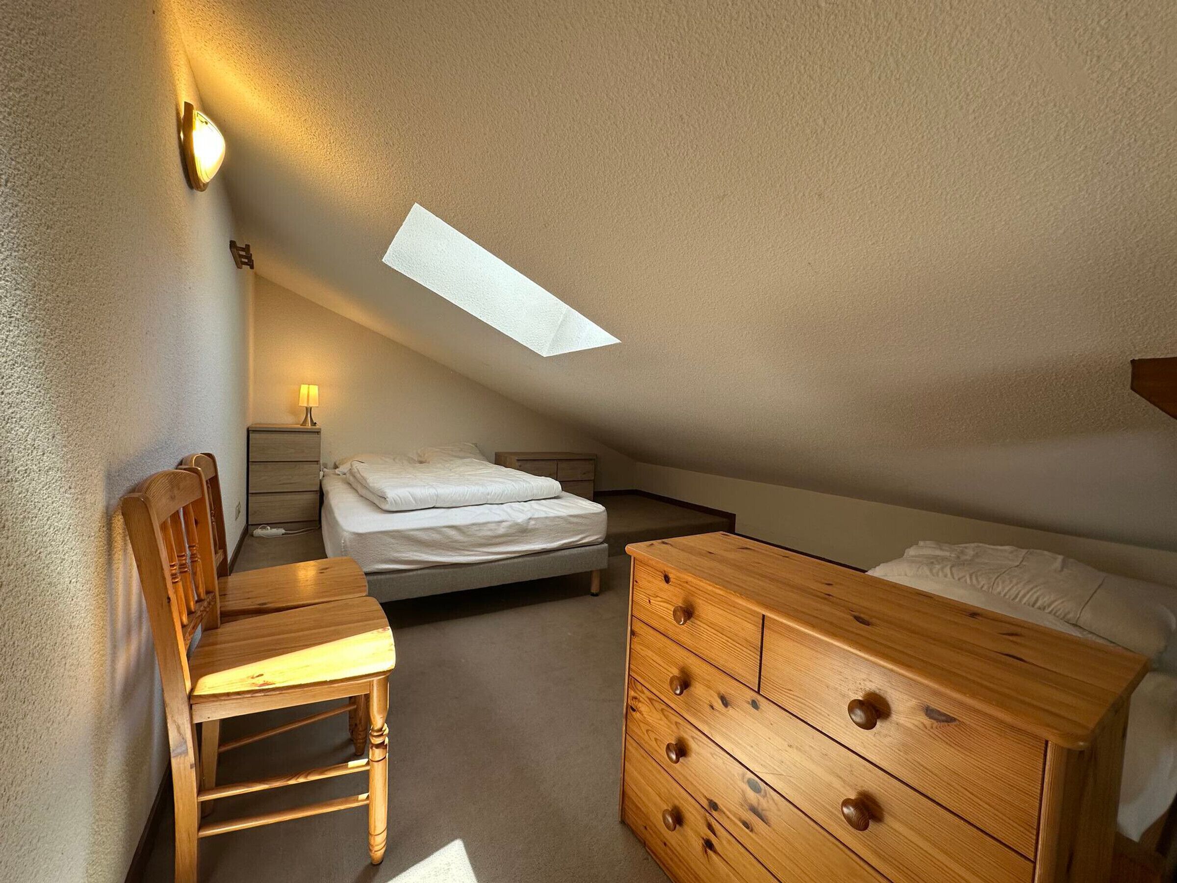 A bedroom with a sloped ceiling, a bed, a wooden dresser, a desk, and a lamp.