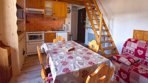 A cozy kitchen with wooden cabinets, a dining table with a patterned tablecloth, and a wooden ladder leading to an upper level. - Charming Duplex Mezzanine in Morillon Village (Morillon Village)