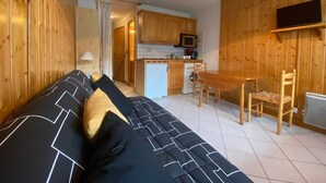 Een gezellige kamer met een bank, een eettafel met stoelen, een keuken met houten kastjes, een wasmachine en een aan de muur bevestigde televisie.