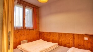 Een kleine kamer met een enkel bed, houten lambrisering, een raam met gordijnen en een plafondlamp.