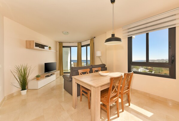 A modern living room with a dining area, a television, and a large window with a view. - 1 bedroom side sea view aparment (Torrox Costa)