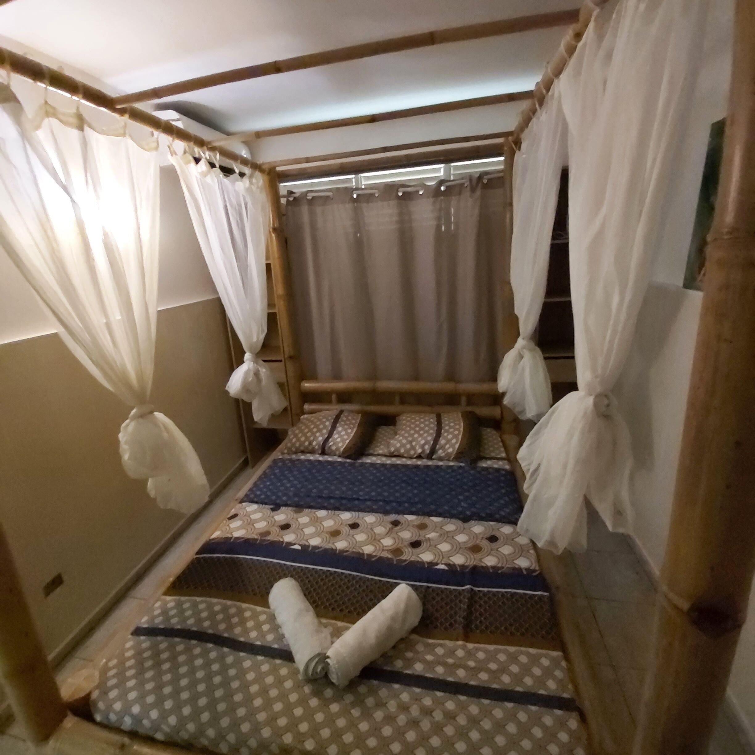 Une petite chambre avec un lit simple, un cadre en bambou, des rideaux blancs et une literie à motifs.
