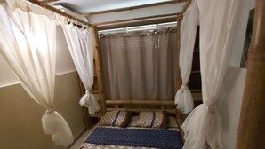 Ein kleines Zimmer mit einem Einzelbett, einem Bambusrahmen, weißen Vorhängen und gemusterten Bettbezügen.