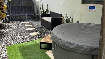 Un jardin avec un jacuzzi, un espace salon couvert et un petit jardin agrémenté de gazon synthétique et de dalles décoratives.