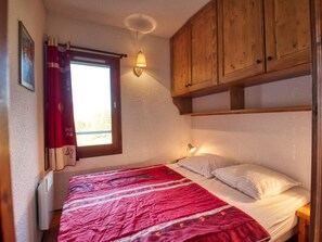 Une chambre à coucher avec un lit, une fenêtre avec des rideaux, un luminaire fixé au mur et des armoires en bois.