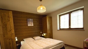 Un dormitorio con cabecera de madera, cama con edredón blanco, dos lámparas de noche, una ventana con cortinas estampadas con corazones y una lámpara colgante.