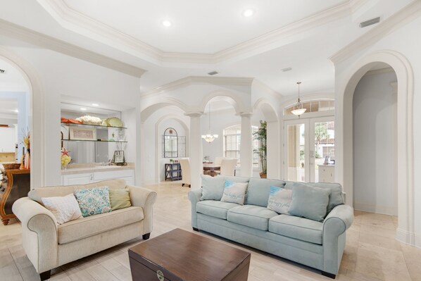 A spacious living room with a beige sofa, a light blue sofa, and a wooden coffee table. There are white walls, arched doorways, and a chandelier.