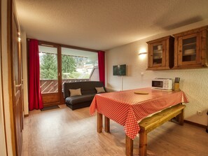 A compact room with a wooden bench, a small table with a red polka dot tablecloth, a black sofa, wooden cabinets, and a view through a large window. - Cozy apartment for 6 near playground (Morillon 1100)