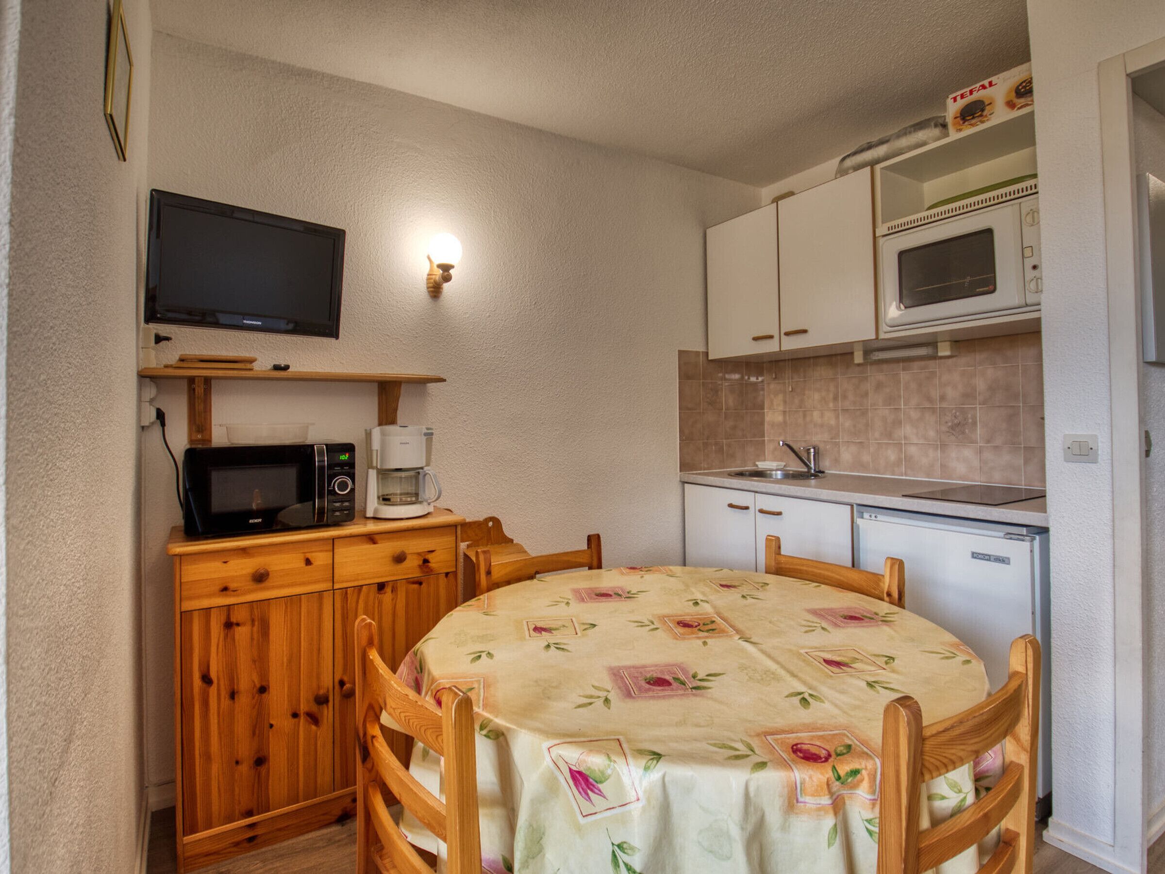 A compact kitchen with a microwave, coffee maker, and a TV on a shelf. There's a dining table with chairs and a cabinet with a TV above it.