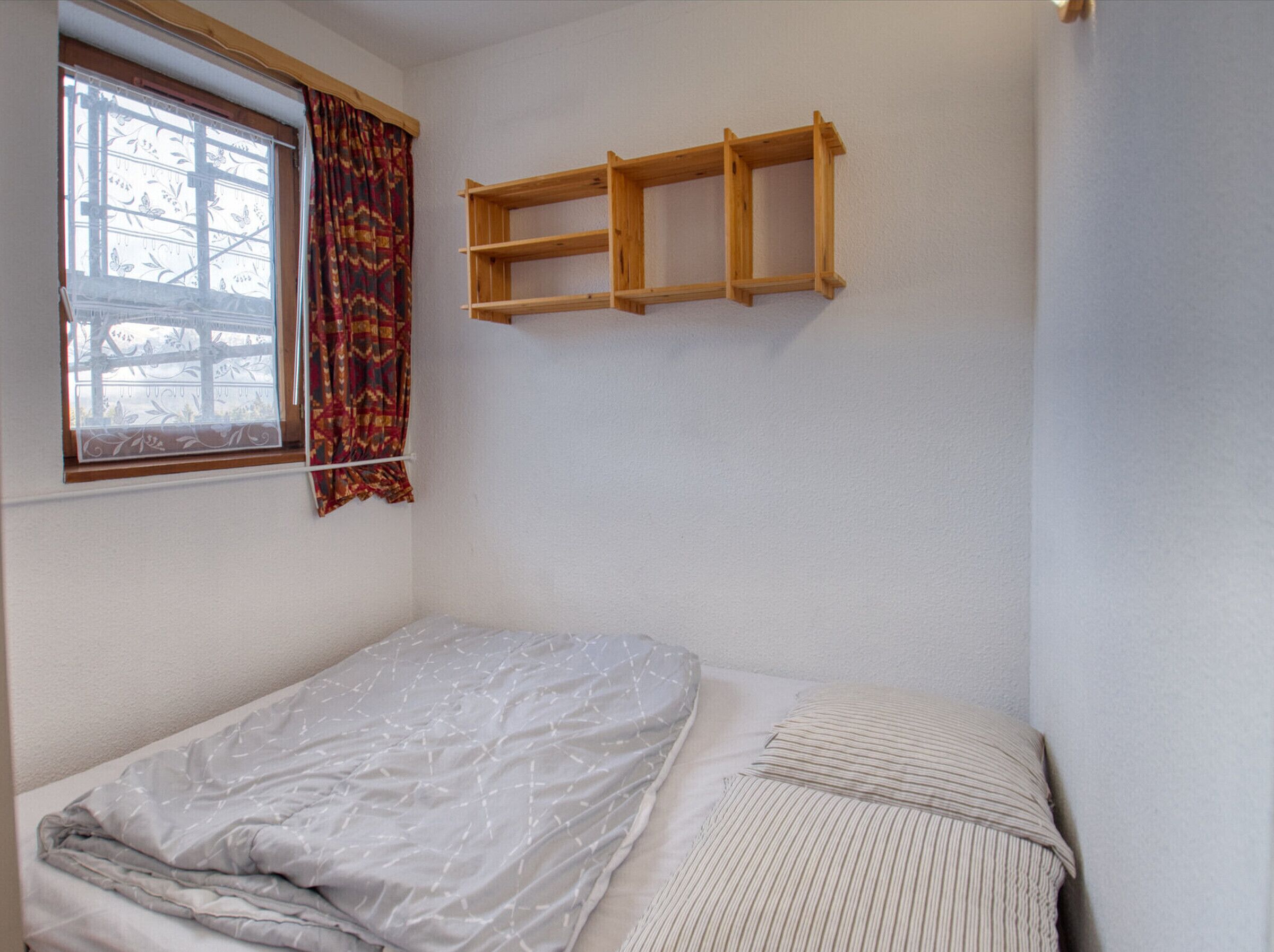 A small, sparsely furnished room with a single bed, a window with patterned curtains, and a wooden shelf unit.