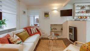 A living room with a white sofa, patterned cushions, a coffee table, a TV, and a bookshelf with various items. - Ground Floor Apartment W/large Enclosed Garden (Crieff)