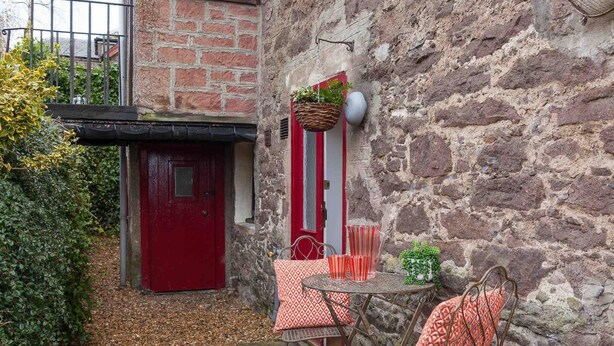 A stone building with a red door, a small table with chairs, and a hanging basket with plants. - Ground Floor Apartment W/large Enclosed Garden (Crieff)