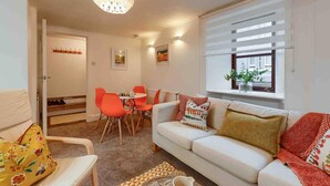 A modern living room with a white sofa, orange chairs, a dining table, and a large window with blinds. - Ground Floor Apartment W/large Enclosed Garden (Crieff)
