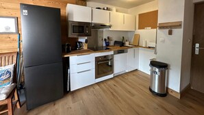 A compact kitchen with white cabinets, a black refrigerator, a microwave, a stove, and a trash bin. - Spacious 3-bedroom duplex at the slopes, parking included (Morillon 1100)