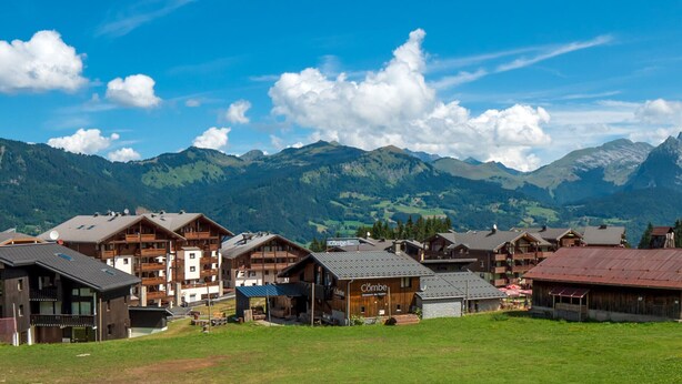 A mountain resort with wooden buildings, a grassy field, and a backdrop of mountains. - Spacious 3-bedroom duplex at the slopes, parking included (Morillon 1100)