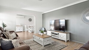 Een moderne woonkamer met een flatscreen televisie, een witte entertainmentkast, een bruine leren bank en een eethoek met witte meubels.