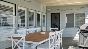Een overdekt terras met een houten tafel en witte stoelen, een barbecue en een deur met horrengaas.