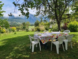 A round table with a floral tablecloth, surrounded by white chairs, set outdoors with a mountain view in the background. - Family home in Morillon, sauna and garden (Morillon Village)