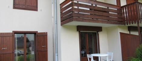 A two-story building with a balcony, a patio with a table and chairs, and wooden shutters on the windows.